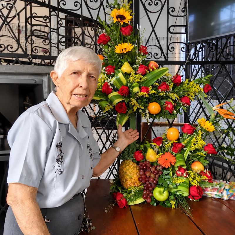 Clarita sonríe junto a un arreglo de rosas, girasoles y frutas, en una fotografía que acompaña el homenaje a su vida.