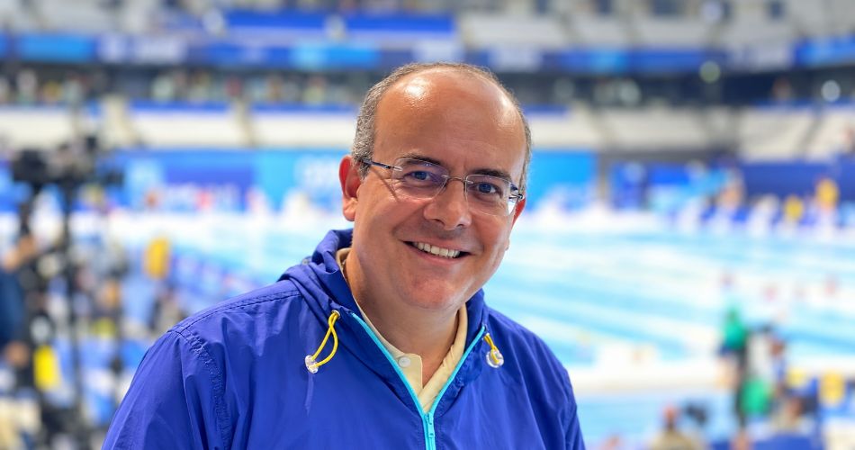 Ricardo Orrego sonríe con gafas y chaqueta azul en un recinto deportivo.