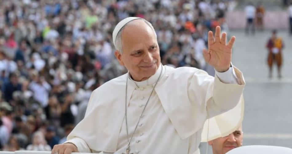 El papa León XIV saluda con la mano levantada detrás de un atril transparente.